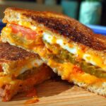 Close-up of a spicy 20-ingredient grilled cheese sandwich, cut in half, with melted cheese, pickles, and tomato.