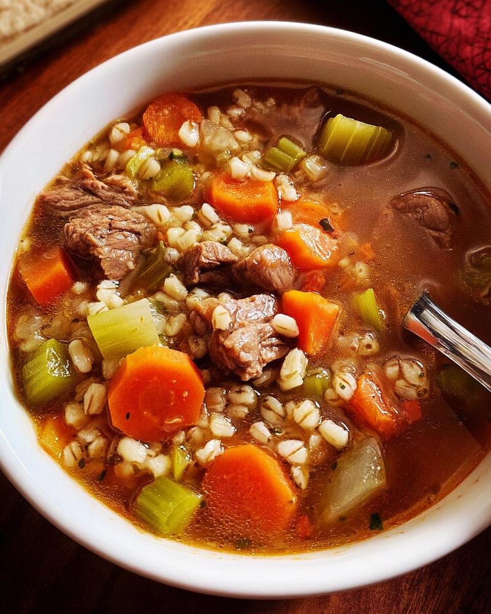 A close-up of a bowl of hearty beef barley soup, packed with tender beef chunks, carrots, celery, and barley.