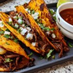 Close-up of three birria tacos, filled with shredded beef and topped with onions and cilantro, served with a side of consommé.