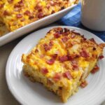 A slice of breakfast casserole with eggs, cheese, hash browns, and bacon on a white plate.