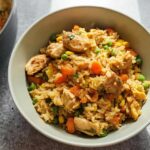 A close-up of a bowl filled with chicken fried rice, featuring chicken pieces, peas, carrots, and corn.