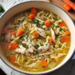 A steaming pot of homemade chicken noodle soup, featuring shredded chicken, wide egg noodles, carrots, and celery, garnished with fresh parsley.