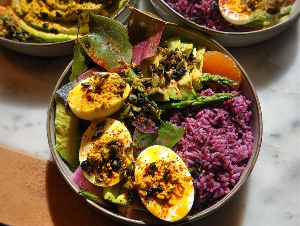 Close-up of a colorful bowl featuring deviled eggs, purple rice, avocado, asparagus, and orange slices.