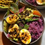 Close-up of a colorful bowl featuring deviled eggs, purple rice, avocado, asparagus, and orange slices.