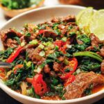 A close-up of a bowl of easy beef and spinach stir-fry with red chili peppers and lime wedges.