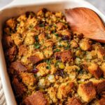 A close-up of a baking dish filled with golden brown stuffing, featuring cubes of bread, dried cranberries, and fresh parsley.