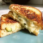 A close-up of a perfectly grilled cheese sandwich, cut in half, showcasing melted cheese and pickle slices.