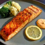 A perfectly grilled salmon fillet served with broccoli, cauliflower, a lemon slice, and sauce. Part of our 12-Ingredient Salmon Recipes.
