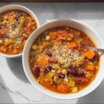 Two white bowls filled with hearty soup recipes, featuring vegetables like carrots, beans, and chickpeas, topped with grated cheese.