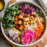 A vibrant and healthy rice bowl with brown rice, kale, roasted sweet potatoes, chickpeas, shredded carrots, red cabbage, and radish slices, drizzled with a yellow dressing.