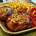 Two perfectly cooked pork chops glazed with sauce and topped with green onions, served with corn and bell peppers.