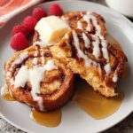 Close-up of restaurant-style cinnamon roll French toast, drizzled with icing and syrup, with raspberries and butter.