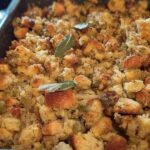 Close-up of a pan filled with freshly baked restaurant-style stuffing, featuring bread cubes, herbs, and sage leaves.