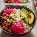 A close-up of a flavorful rice bowl recipe meal prep, featuring rice, ground meat, black beans, avocado, cheese, and pickled onions.