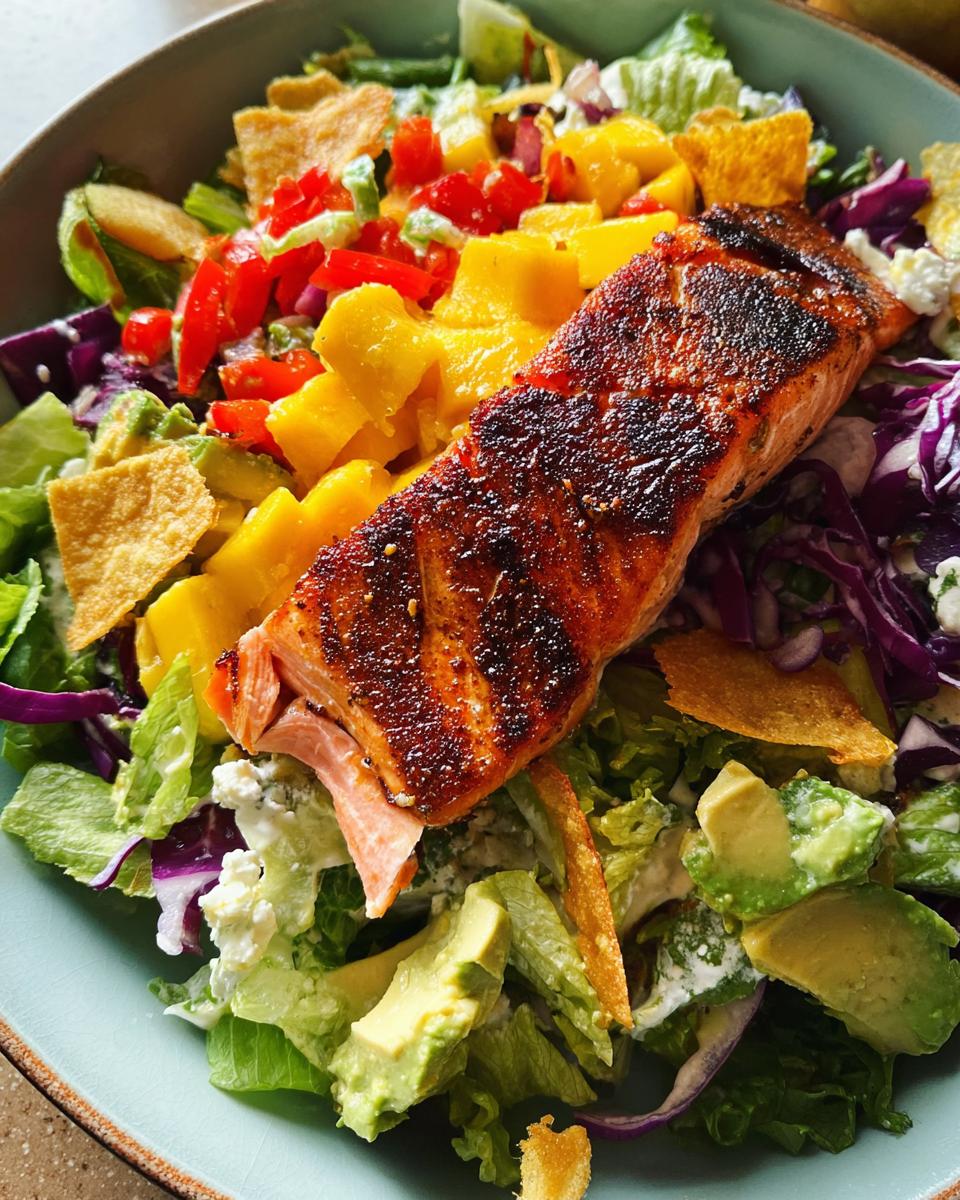 A vibrant salad topped with a perfectly cooked salmon fillet, featuring mango, avocado, red cabbage, and crispy tortilla strips.
