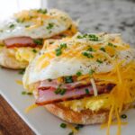 Close-up of two delicious sandwich ideas recipes with ham, scrambled eggs, a fried egg, and shredded cheese on an English muffin.
