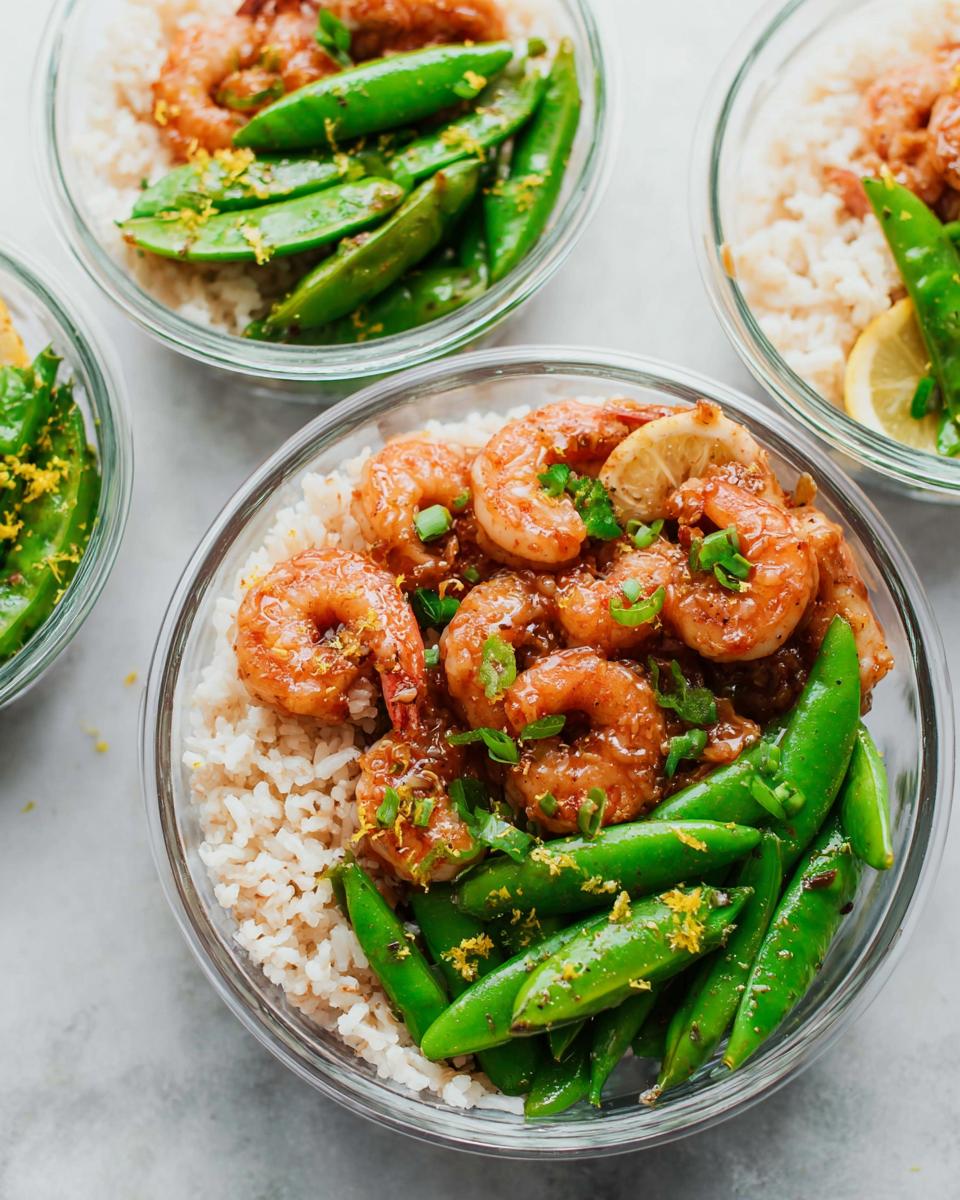 Three glass bowls filled with shrimp recipes meal prep: brown rice, glazed shrimp, and snap peas, garnished with lemon zest.
