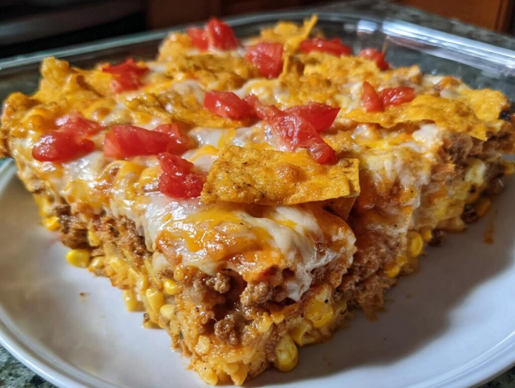 A hearty slice of Taco Tuesday casserole featuring layers of ground meat, corn, cheese, and crushed tortilla chips, topped with diced tomatoes.