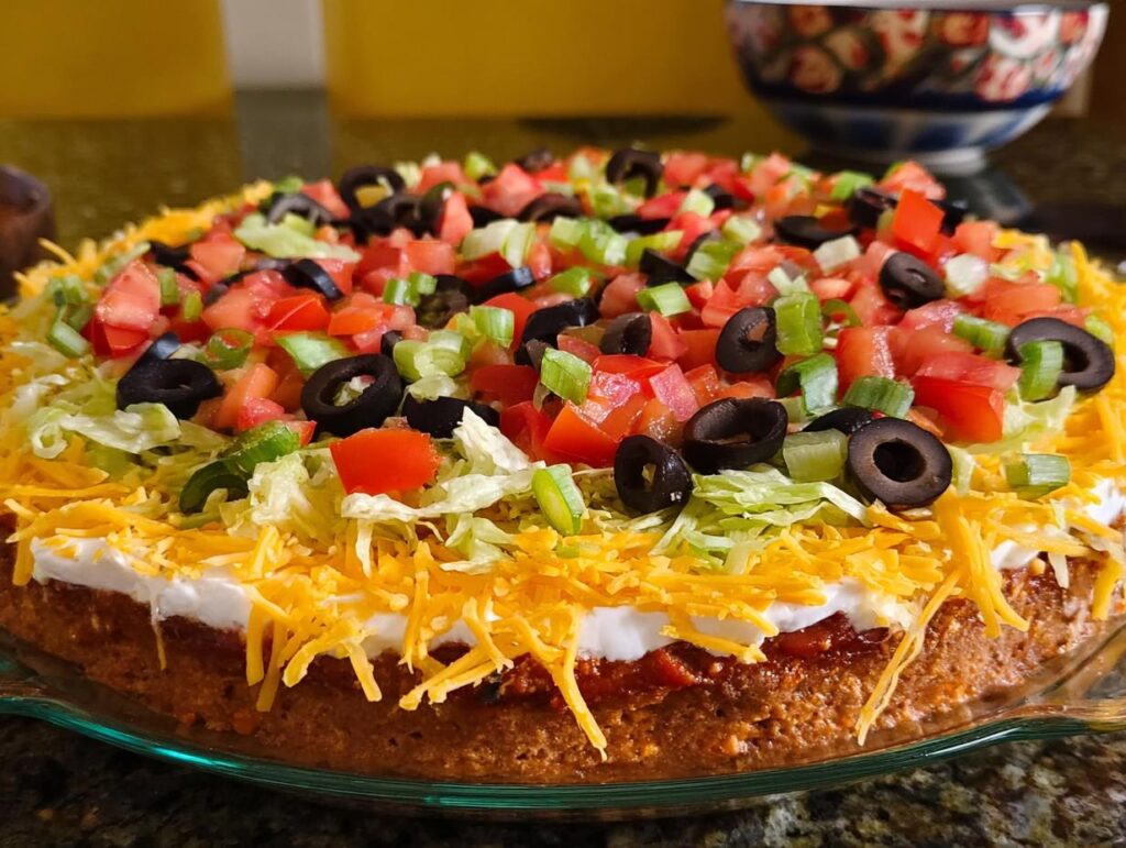 A delicious Taco Tuesday recipe featuring a layered taco casserole topped with shredded cheese, lettuce, tomatoes, olives, and green onions.