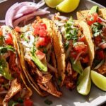 A plate of freshly prepared chicken tacos for Taco Tuesday Meal Prep, topped with avocado, tomato, onion, and cilantro, served with lime wedges.