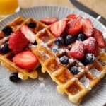 Two golden waffles topped with fresh strawberries, blueberries, and powdered sugar. A perfect breakfast idea.