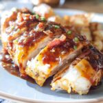 A close-up of sliced Cowboy BBQ Bacon Cheddar Chicken, drizzled with sauce and topped with bacon bits and chives.