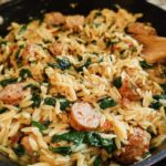 Close-up of creamy chicken sausage orzo with spinach in a skillet, being stirred with a wooden spoon.