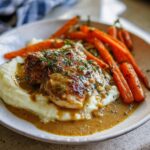 A plate of creamy herb chicken served with mashed potatoes and glazed carrots, topped with fresh herbs.