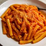 A close-up of a bowl of creamy tomato garlic pasta, topped with grated cheese and black pepper.