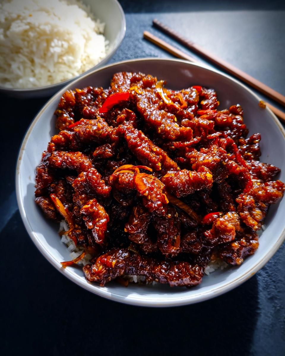 A bowl of fluffy white rice topped with glistening, saucy Crispy Chilli Beef and sliced red chilies.