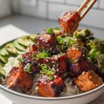 A close-up of a Crispy Salmon and Rice Bowl, featuring glazed salmon cubes, rice, sliced cucumbers, and broccoli florets, with chopsticks lifting a piece of salmon.