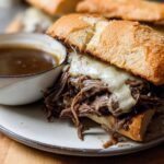 A close-up of a Crockpot French Dip Sandwich overflowing with tender shredded beef and melted provolone cheese, served with a side of au jus.