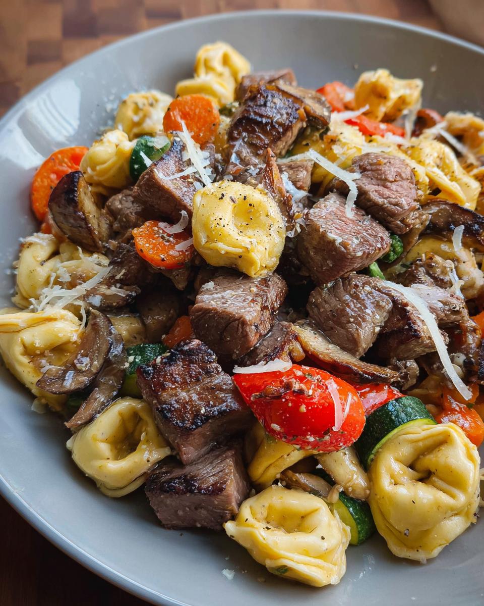 A close-up of a bowl of Garlic Steak Tortellini, featuring tender steak pieces, tortellini pasta, carrots, zucchini, and mushrooms.