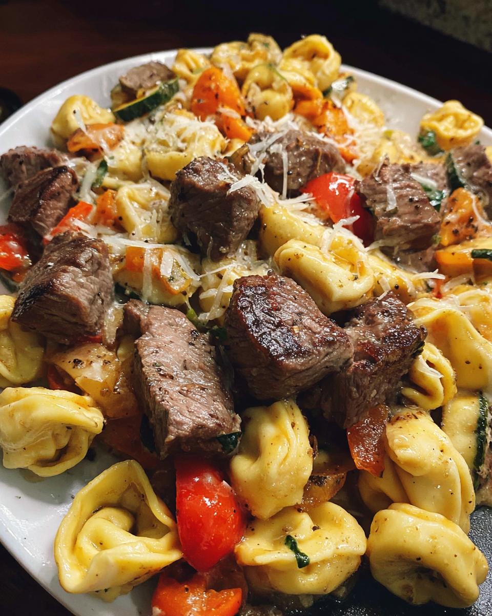 A close-up of Garlic Steak Tortellini pasta dish with chunks of steak, tortellini, tomatoes, and zucchini, topped with grated cheese.