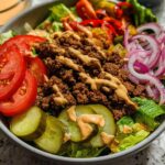A delicious High-Protein Cheeseburger Bowl filled with seasoned ground beef, lettuce, tomatoes, pickles, and red onion, drizzled with sauce.