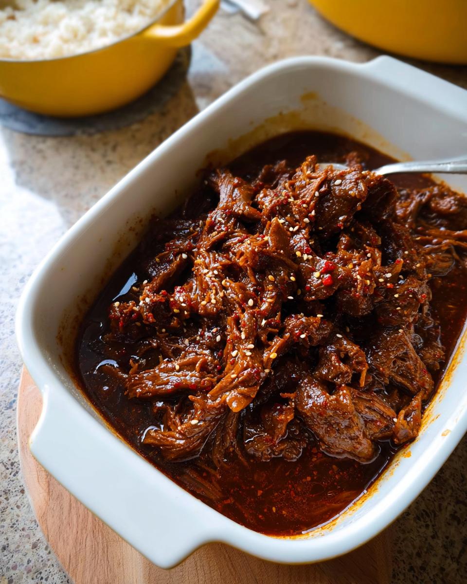 A close-up of tender Korean Style Pot Roast, shredded and coated in a rich, dark sauce, sprinkled with sesame seeds.