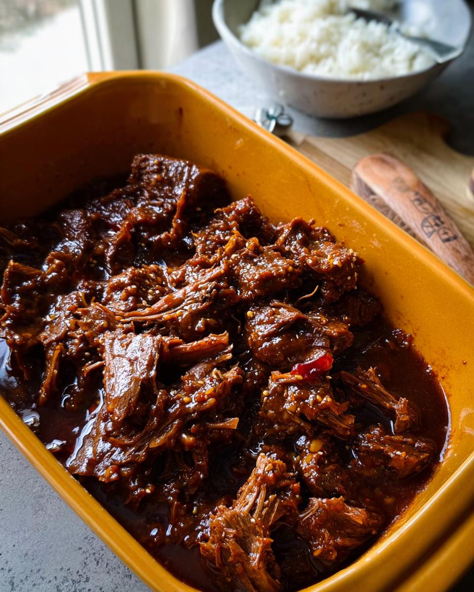 Close-up of tender Korean Style Pot Roast shredded and coated in a rich, dark sauce, served in a yellow dish.