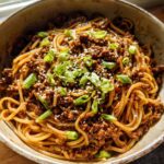 A close-up of a rustic bowl filled with Mongolian Ground Beef Noodles, garnished with green onions and sesame seeds.
