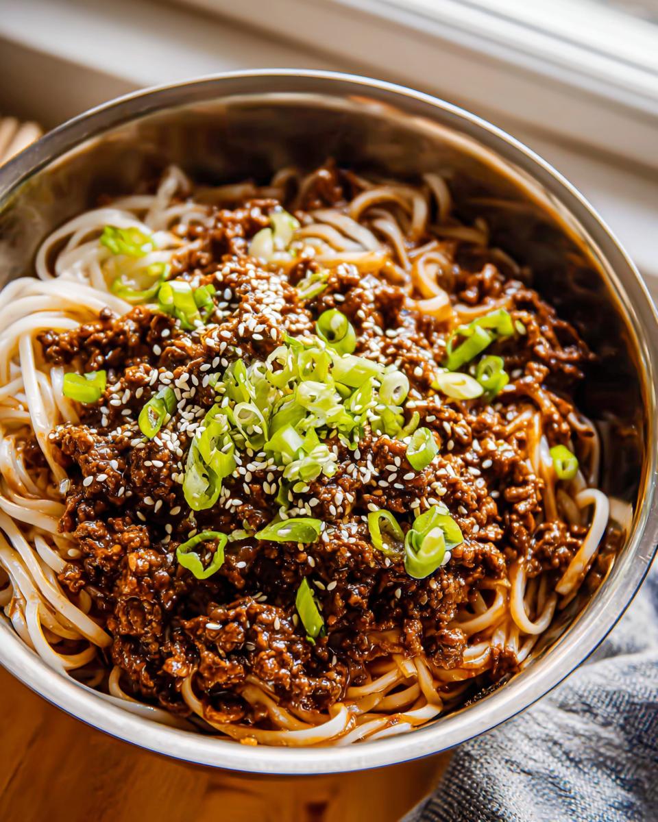 A close-up overhead view of a bowl filled with Mongolian Ground Beef Noodles, topped with sesame seeds and chopped green onions.