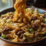 A close-up of a bowl of Potsticker Noodle Bowl with Pork & Cabbage, with noodles being lifted by chopsticks.