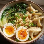 A close-up of a Savory Dumpling Ramen Bowl with soft-boiled eggs, bok choy, and chili flakes.