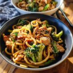 A close-up of a bowl filled with Spicy Garlic Chicken and Broccoli Noodle Bowls, garnished with sesame seeds.