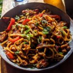 A close-up overhead view of a bowl filled with delicious Sticky Garlic Chicken Noodles, garnished with green onions and red bell peppers.