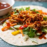 A delicious Sweet Chili Chicken Wrap filled with shredded chicken, lettuce, cucumber, carrots, and bell peppers, drizzled with sauce.