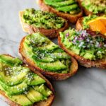 A variety of delicious avocado toast preparations on toasted bread, showcasing sliced avocado, mashed avocado with red onion, and orange slices.
