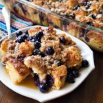 A delicious slice of Best Blueberry French Toast Casserole, topped with streusel and fresh blueberries, served with syrup.