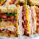A close-up of a stacked Breakfast Club Sandwich with layers of turkey, bacon, cheese, lettuce, and tomato.