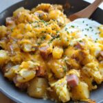 A close-up of a Cheesy Potato Egg Scramble in a skillet, topped with melted cheddar cheese and chives.
