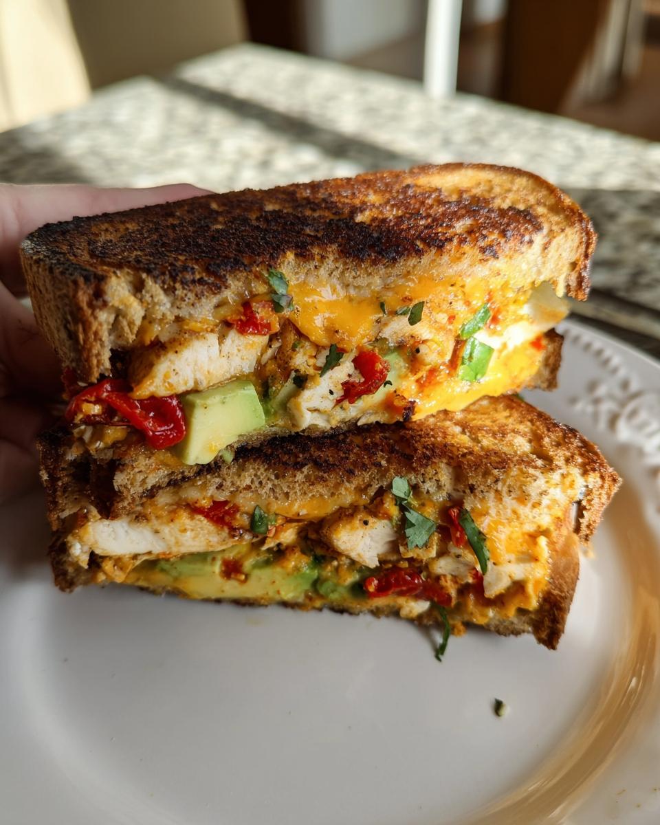 A close-up of a halved CHICKEN AVOCADO MELT SANDWICH on a white plate, showing layers of grilled chicken, avocado, cheese, and roasted red peppers.