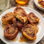 A plate of golden brown Cinnamon Roll French Toast Bites, sprinkled with cinnamon sugar and drizzled with syrup.